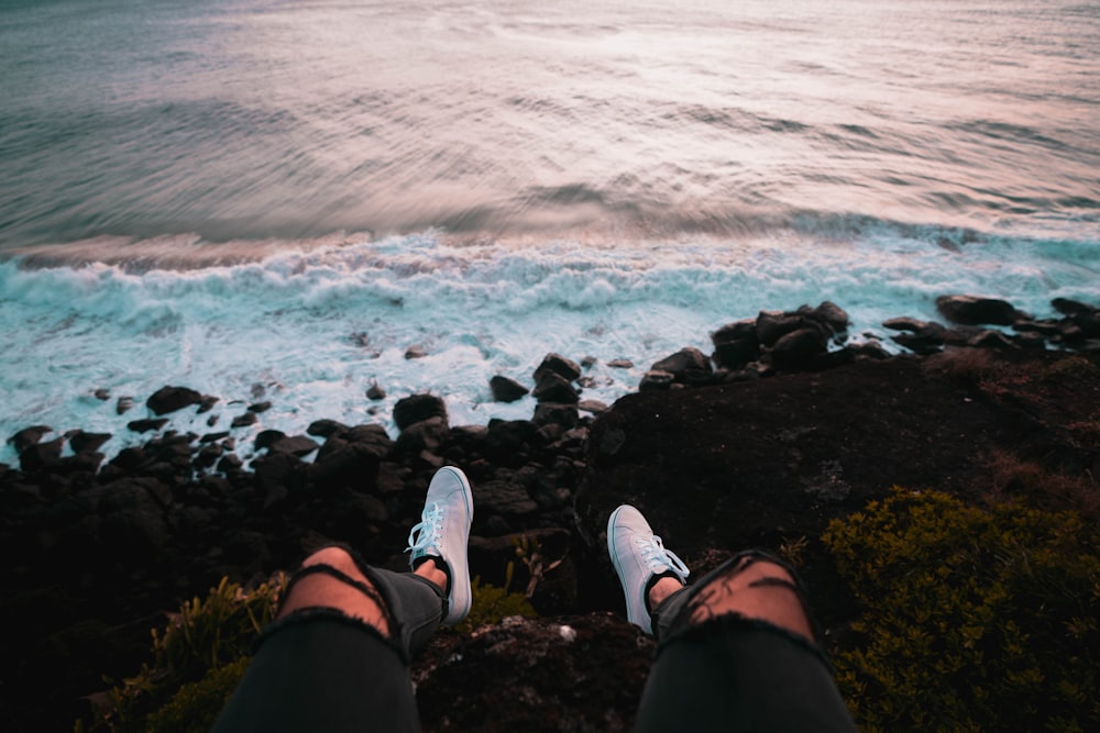 Person in schwarzen Shorts und weißen Turnschuhen, die tagsüber auf dem Felsen am Meer sitzt