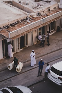 A street scene featuring several people outside a furniture store with a sign that reads 'AL KHAJA FURNITURE'. One man is carrying two rolled-up carpets, another man in a white robe is standing nearby, and three other individuals are engaged in conversation close to the store entrance. Several cars are parked alongside the pavement.