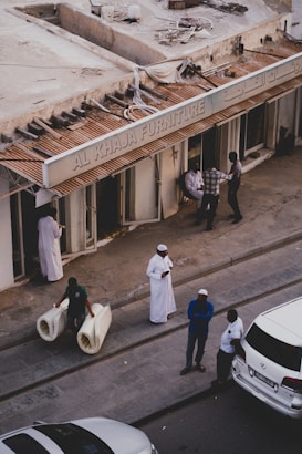 A street scene featuring several people outside a furniture store with a sign that reads 'AL KHAJA FURNITURE'. One man is carrying two rolled-up carpets, another man in a white robe is standing nearby, and three other individuals are engaged in conversation close to the store entrance. Several cars are parked alongside the pavement.