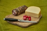 A wooden cutting board holds a block of cheese, several red radishes, a piece of cured meat, and a black-handled knife against a green background.