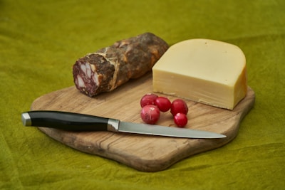 A wooden cutting board holds a block of cheese, several red radishes, a piece of cured meat, and a black-handled knife against a green background.