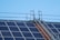 Solar panels are installed on a rooftop along with a metal walkway or ladder structure for maintenance access. The sky in the background is clear and blue.