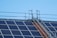 Solar panels are installed on a rooftop along with a metal walkway or ladder structure for maintenance access. The sky in the background is clear and blue.