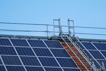 Solar panels are installed on a rooftop along with a metal walkway or ladder structure for maintenance access. The sky in the background is clear and blue.