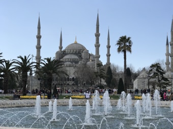 A grand mosque with multiple minarets is set against a clear blue sky. In the foreground, an ornamental fountain sprays water, surrounded by palm trees and lush greenery. People are walking and gathering around the area, adding vibrancy to the scene.
