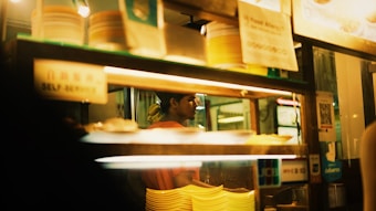The image depicts a food stall or restaurant interior with shelves holding stacks of yellow and white plastic plates. Warm lighting creates a cozy atmosphere. A person is seen in the background, partially obscured by the shelves, and various signs and stickers are visible, including one with the words 'SELF SERVICE'. There is a reflection on the glass, adding depth to the scene.