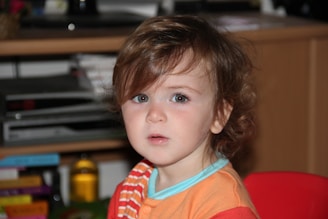 A young child with light brown hair and expressive eyes sits in front of a blurred background that appears to be a living room with shelves and books. The child is wearing a multicolored striped shirt and is looking directly at the camera with a curious expression.