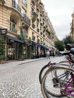 A European city street lined with elegant, historic buildings featuring ornate balconies and window boxes filled with greenery. The street is paved with cobblestones, and several bicycles are parked at the edge. People are strolling along the sidewalk, creating a lively yet calm atmosphere.