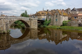 A stone bridge spans over a calm river, reflecting in the water below. On the riverbank, historic buildings with various colored roofs and greenery are visible, creating a picturesque scene reminiscent of a quaint European village.
