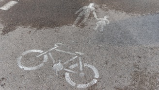 A wet road surface displays white painted markings, including a bicycle symbol and an icon representing an adult holding a child's hand. The dark patches of moisture create a slight contrast with the gray asphalt, highlighting the road symbols clearly.