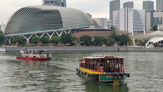 A river scene with two boats, one colorful and wooden in the foreground and another boat with people in the midground. The backdrop displays modern architecture, including a uniquely designed building with a rounded, spiky exterior resembling a durian fruit. Trees line the shore, adding greenery to the urban landscape.