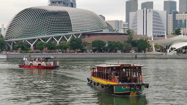 A river scene with two boats, one colorful and wooden in the foreground and another boat with people in the midground. The backdrop displays modern architecture, including a uniquely designed building with a rounded, spiky exterior resembling a durian fruit. Trees line the shore, adding greenery to the urban landscape.