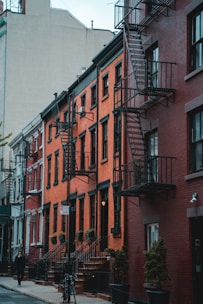 Rows of historic brick apartment buildings with metal fire escapes line a city street. The buildings are closely packed, with large windows and black railings. Small trees in planters and a bicycle are visible on the sidewalk. A single person walks along the street, and a 'No Parking Anytime' sign is posted.