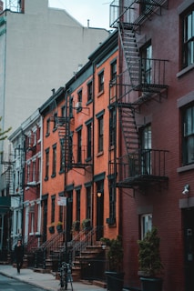 Rows of historic brick apartment buildings with metal fire escapes line a city street. The buildings are closely packed, with large windows and black railings. Small trees in planters and a bicycle are visible on the sidewalk. A single person walks along the street, and a 'No Parking Anytime' sign is posted.