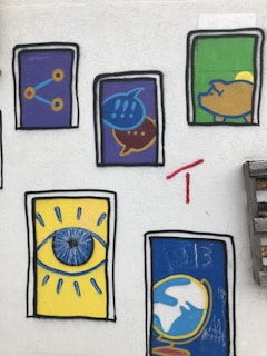 A wall decorated with colorful street art featuring six framed graphics. The top left graphic shows a simple network diagram with three interconnected circles set on a purple background. Next to it, there's a design with two overlapping speech bubbles, one blue with exclamation marks and the other yellow with dots, also on a purple background. The next frame presents a stylized yellow pig on a green background. Below, the bottom left has an eye on a yellow background with blue highlights, while the adjacent illustration is a globe on a blue background outlined in yellow. The scene includes vibrant, bold colors and basic geometric shapes.