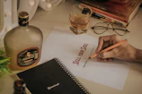 A hand holding a pencil appears to be signing a paper with the words 'SELL YOUR SOUL' printed on it. Nearby are a glass filled with a drink, a bottle with a label, a pair of glasses, a black notebook, and some books stacked on top of each other. The setting suggests a casual workspace or study area.