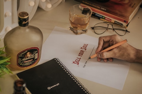 A hand holding a pencil appears to be signing a paper with the words 'SELL YOUR SOUL' printed on it. Nearby are a glass filled with a drink, a bottle with a label, a pair of glasses, a black notebook, and some books stacked on top of each other. The setting suggests a casual workspace or study area.