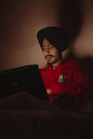 A person wearing a black turban and red hoodie is using a laptop in a dimly lit room. The light from the laptop screen illuminates the face, creating a focused and introspective atmosphere.