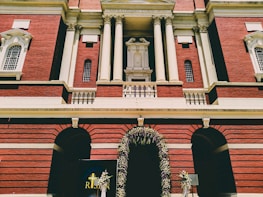 A grand building with red brick architecture, featuring intricate white columns and decorative elements. There is a prominent arched entrance adorned with a floral arrangement. Beneath the archway, a sign with the word 'RISEN' and a cross symbol is visible.