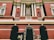 A grand building with red brick architecture, featuring intricate white columns and decorative elements. There is a prominent arched entrance adorned with a floral arrangement. Beneath the archway, a sign with the word 'RISEN' and a cross symbol is visible.