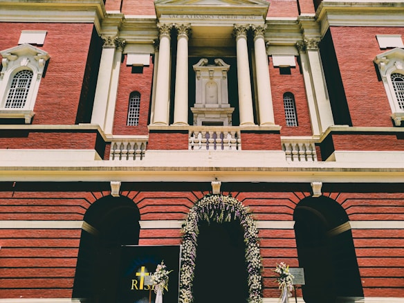 A grand building with red brick architecture, featuring intricate white columns and decorative elements. There is a prominent arched entrance adorned with a floral arrangement. Beneath the archway, a sign with the word 'RISEN' and a cross symbol is visible.