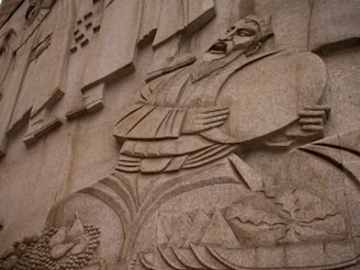 A detailed stone carving depicts a historical or cultural figure in traditional attire, holding a large circular object near his chest. The figure has a long beard and an expressive face. The background includes geometric patterns and a collection of carved elements resembling food items or offerings, suggesting themes of abundance or prosperity.