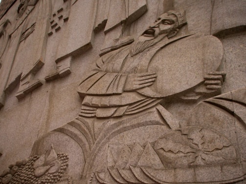 A detailed stone carving depicts a historical or cultural figure in traditional attire, holding a large circular object near his chest. The figure has a long beard and an expressive face. The background includes geometric patterns and a collection of carved elements resembling food items or offerings, suggesting themes of abundance or prosperity.