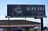 A large billboard displays an advertisement for the film 'Bird Box' featuring an image of two individuals wearing blindfolds. The scene is dramatic and tense, indicative of the film's survival theme. The names of the actors, director, and streaming platform are prominently featured.