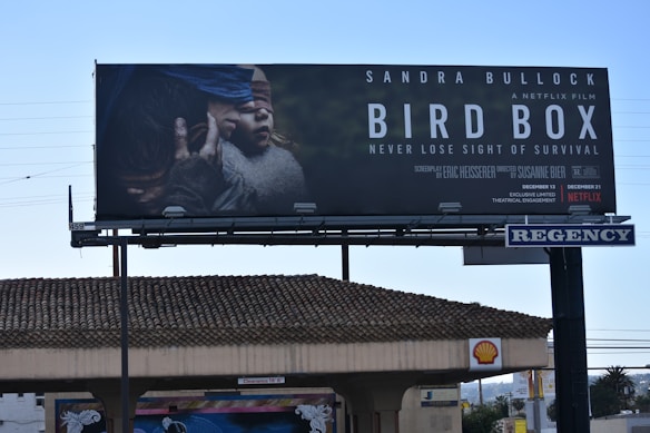 A large billboard displays an advertisement for the film 'Bird Box' featuring an image of two individuals wearing blindfolds. The scene is dramatic and tense, indicative of the film's survival theme. The names of the actors, director, and streaming platform are prominently featured.
