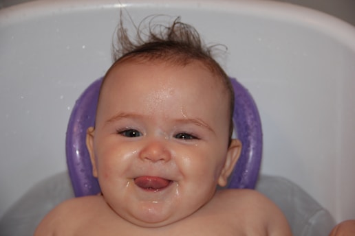 A smiling baby with wet hair, seated in a bathtub with a purple bath support or cushion. The baby's tongue is sticking out slightly, and water droplets are visible on the skin, conveying a sense of playfulness and joy during bath time.
