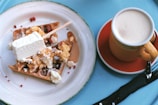A plate of waffles topped with various ingredients and a stick of ice cream or dessert, paired with a cup of frothy coffee. The table is covered with a blue tablecloth.