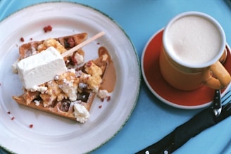 A plate of waffles topped with various ingredients and a stick of ice cream or dessert, paired with a cup of frothy coffee. The table is covered with a blue tablecloth.