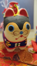 A colorful hand-painted figurine of a cat features prominent blue and red facial details, large eyes, and a small red mouth. The cat is adorned with intricate floral patterns in yellow, white, and green on its body. It rests on a vibrant red and orange patterned fabric.