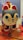 A colorful hand-painted figurine of a cat features prominent blue and red facial details, large eyes, and a small red mouth. The cat is adorned with intricate floral patterns in yellow, white, and green on its body. It rests on a vibrant red and orange patterned fabric.