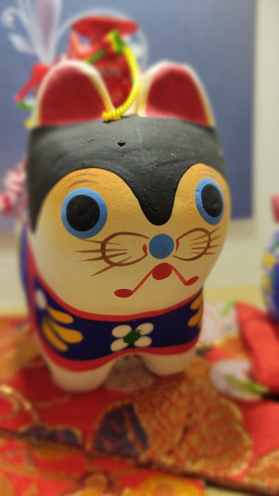A colorful hand-painted figurine of a cat features prominent blue and red facial details, large eyes, and a small red mouth. The cat is adorned with intricate floral patterns in yellow, white, and green on its body. It rests on a vibrant red and orange patterned fabric.
