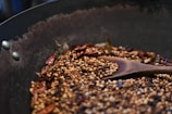 A metal pan contains a variety of toasted spices including coriander seeds, peppercorns, and dried red chilies. A wooden spoon is resting among the spices, all of which are lightly browned, suggesting they are being roasted. The surface of the pan appears seasoned from previous use.