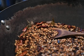 A metal pan contains a variety of toasted spices including coriander seeds, peppercorns, and dried red chilies. A wooden spoon is resting among the spices, all of which are lightly browned, suggesting they are being roasted. The surface of the pan appears seasoned from previous use.