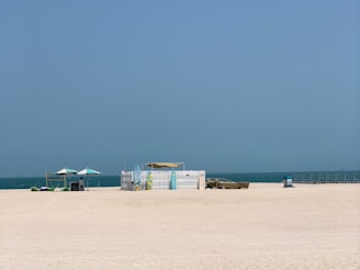 A sandy beach stretches in the foreground with a clear, blue sky above. In the middle distance, there is a white picket fence structure surrounded by beach chairs and umbrellas. A few colorful inflatable floats are scattered nearby. The ocean is calm and lies beyond the beach setup.