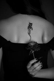 A person with long dark hair is seen from the back, revealing a stylized tattoo on their upper back. They are wearing a dark off-the-shoulder top and holding a single rose in one hand. The image is black and white, giving it a classic and mysterious feel.