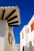 Whitewashed Mediterranean-style buildings with distinctive, dark wooden beams contrast against a bright blue sky. The structures feature minimalistic windows and are bathed in sunlight, highlighting the architectural design.