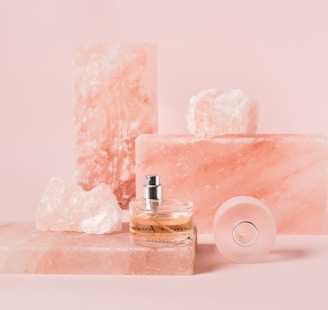 A collection of pink Himalayan salt blocks and crystals arranged with a clear glass perfume bottle, which has a silver spray nozzle. The background and overall color tone is a soft pink, contributing to a calm and elegant aesthetic.