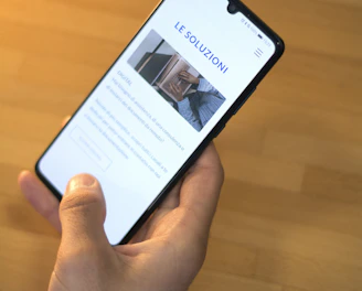 A hand is holding a smartphone displaying a website with the header 'LE SOLUZIONI'. The smartphone screen shows a section with an image of a person and text underneath. The background is a wooden table surface.