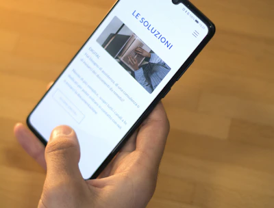 A hand is holding a smartphone displaying a website with the header 'LE SOLUZIONI'. The smartphone screen shows a section with an image of a person and text underneath. The background is a wooden table surface.