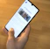 A hand is holding a smartphone displaying a website with the header 'LE SOLUZIONI'. The smartphone screen shows a section with an image of a person and text underneath. The background is a wooden table surface.