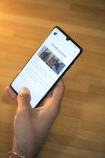 A hand is holding a smartphone displaying a website with the header ''. The smartphone screen shows a section with an image of a person and text underneath. The background is a wooden table surface.