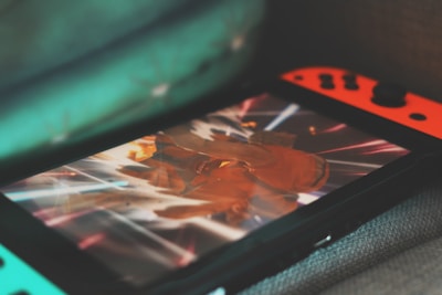 A portable gaming console with neon red and blue controllers displays an action-packed scene from a video game. The screen is focused on a character surrounded by bright, dynamic light effects.