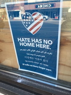 A blue poster with a heart shape containing the American flag design. The text 'Hate Has No Home Here' is prominently displayed in English, followed by translations in several other languages. The poster is placed on a window, with reflections of the street and a cinema across the road visible.