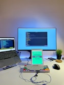 A modern workspace featuring a large monitor, a laptop, an iPad displaying a book titled 'Mastering Vim Quickly,' a mechanical keyboard with colorful keycaps, a wireless mouse, a potted plant, a smartphone, pens, a notebook, and earphones on a white desk. The monitor and the laptop both display code editors.