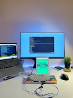 A modern workspace featuring a large monitor, a laptop, an iPad displaying a book titled 'Mastering Vim Quickly,' a mechanical keyboard with colorful keycaps, a wireless mouse, a potted plant, a smartphone, pens, a notebook, and earphones on a white desk. The monitor and the laptop both display code editors.