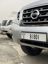 A close-up view of several parked Nissan cars, focusing on the front grille and license plate of the nearest vehicle, which has a Dubai registration number.